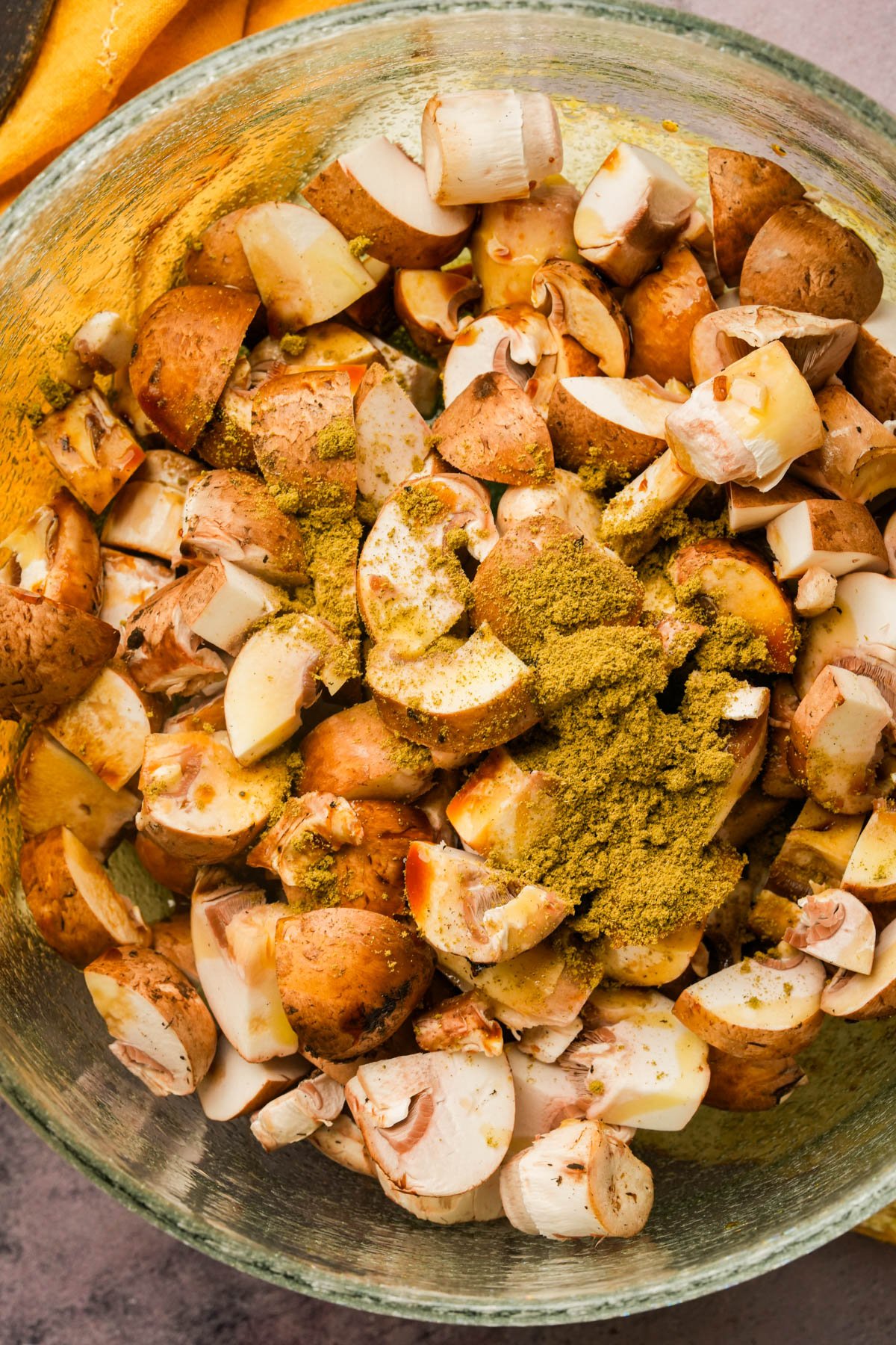 A glass bowl filled with chopped brown mushrooms topped with poultry seasoning, ready to be mixed for cooking.