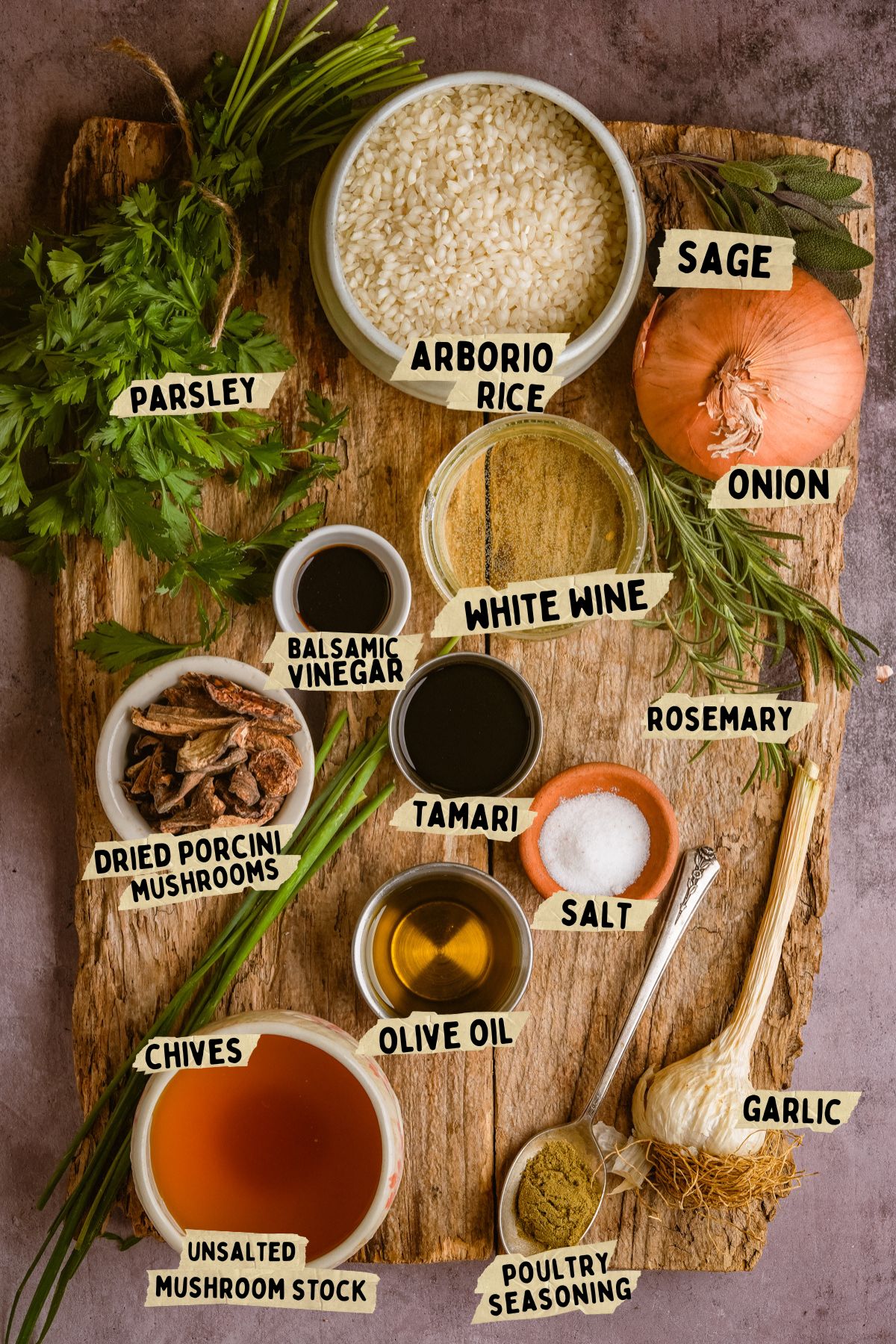 A wooden board displays labeled ingredients for a mushroom risotto recipe: parsley, sage, arborio rice, onion, white wine, rosemary, garlic, poultry seasoning, salt, olive oil, tamari, balsamic vinegar, dried porcini mushrooms, chives, mushroom stock.
