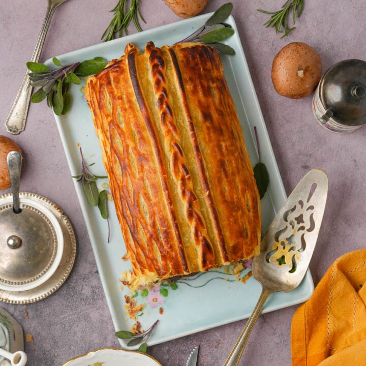 Golden-brown vegan mushroom Wellington on a rectangular plate, garnished with fresh herbs. Surrounded by mushrooms, silverware, a pepper shaker, and a folded orange napkin on a light purple surface.