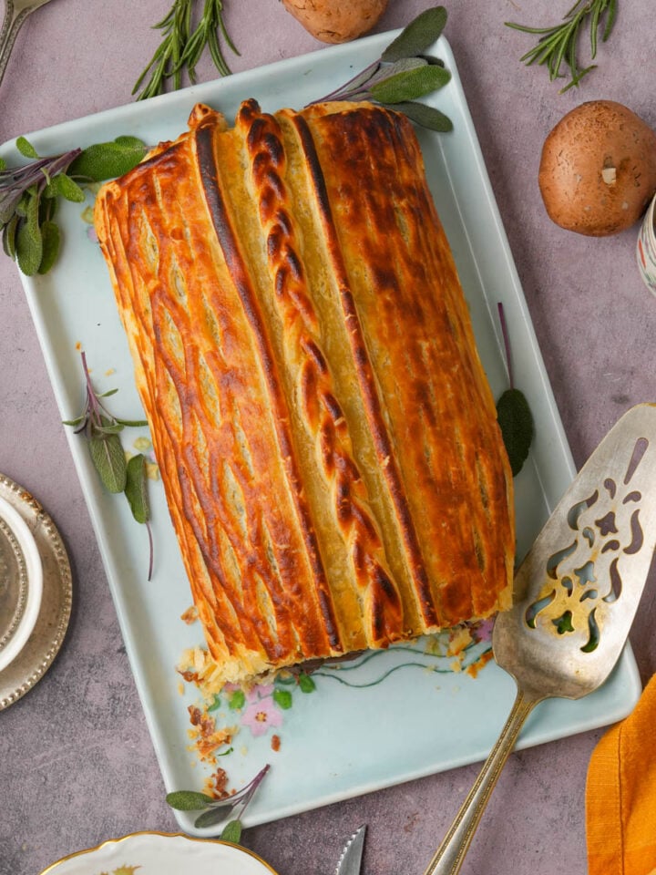 Golden-brown vegan mushroom Wellington on a rectangular plate, garnished with fresh herbs. Surrounded by mushrooms, silverware, a pepper shaker, and a folded orange napkin on a light purple surface.