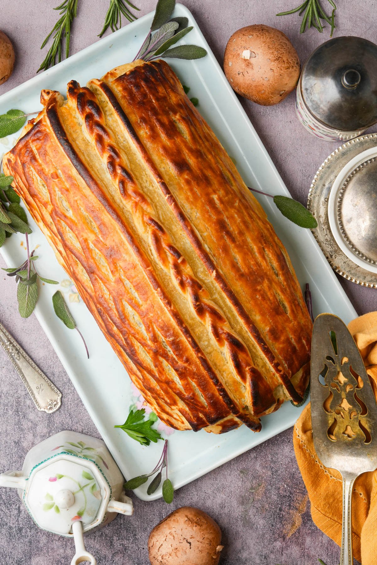 A golden-brown baked vegan mushroom Wellington on a rectangular platter, garnished with fresh herbs. Whole mushrooms, vintage silverware, and a teapot are arranged around it on a light textured surface.