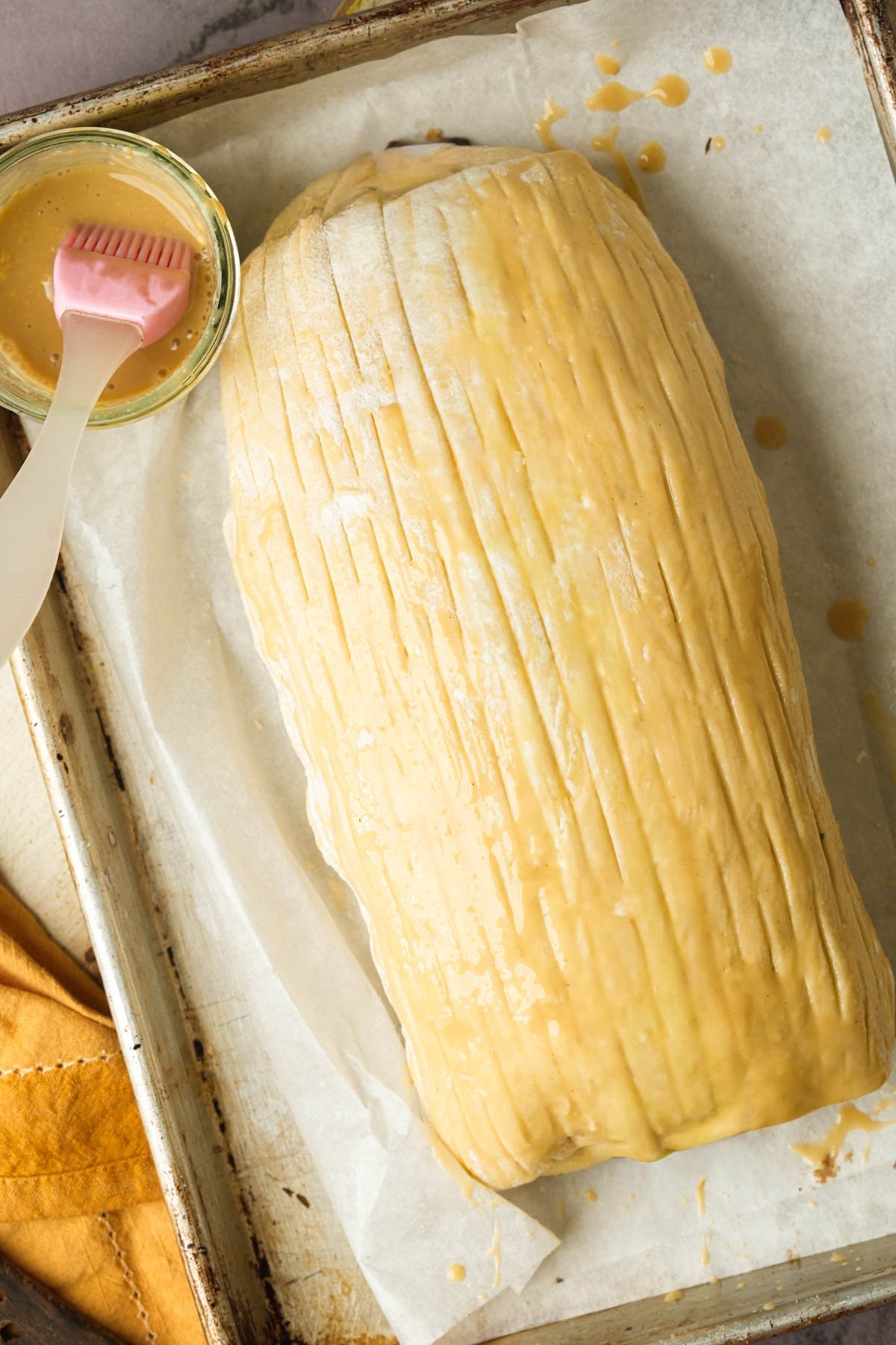 A large, unbaked vegan mushroom Wellington loaf covered in a second sheet of scored dough rests on parchment paper in a baking tray. A pastry brush and a small bowl of egg wash sit beside it, ready for further glazing.