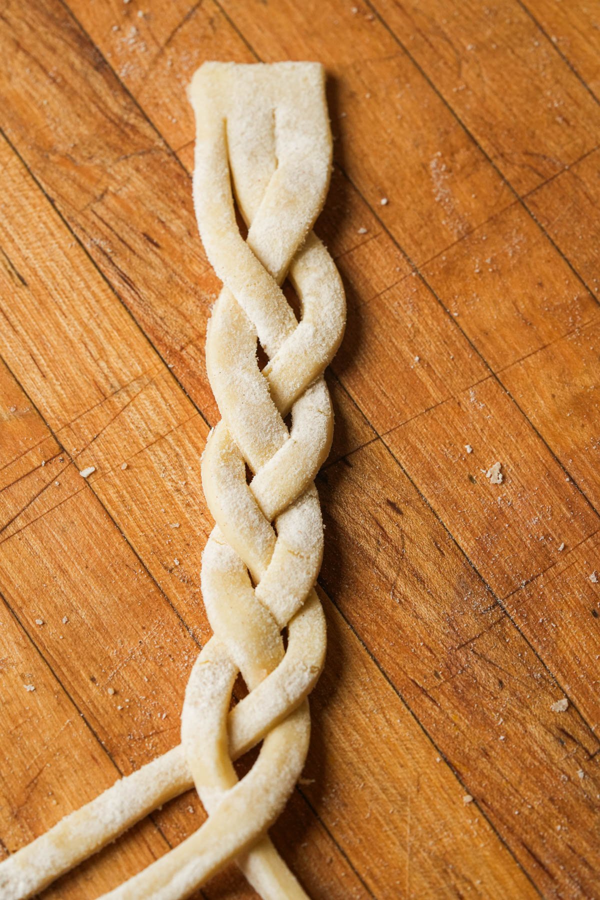 Three strips of dough are braided together on a wooden surface, with flour lightly dusted on the dough and some scattered across the table.