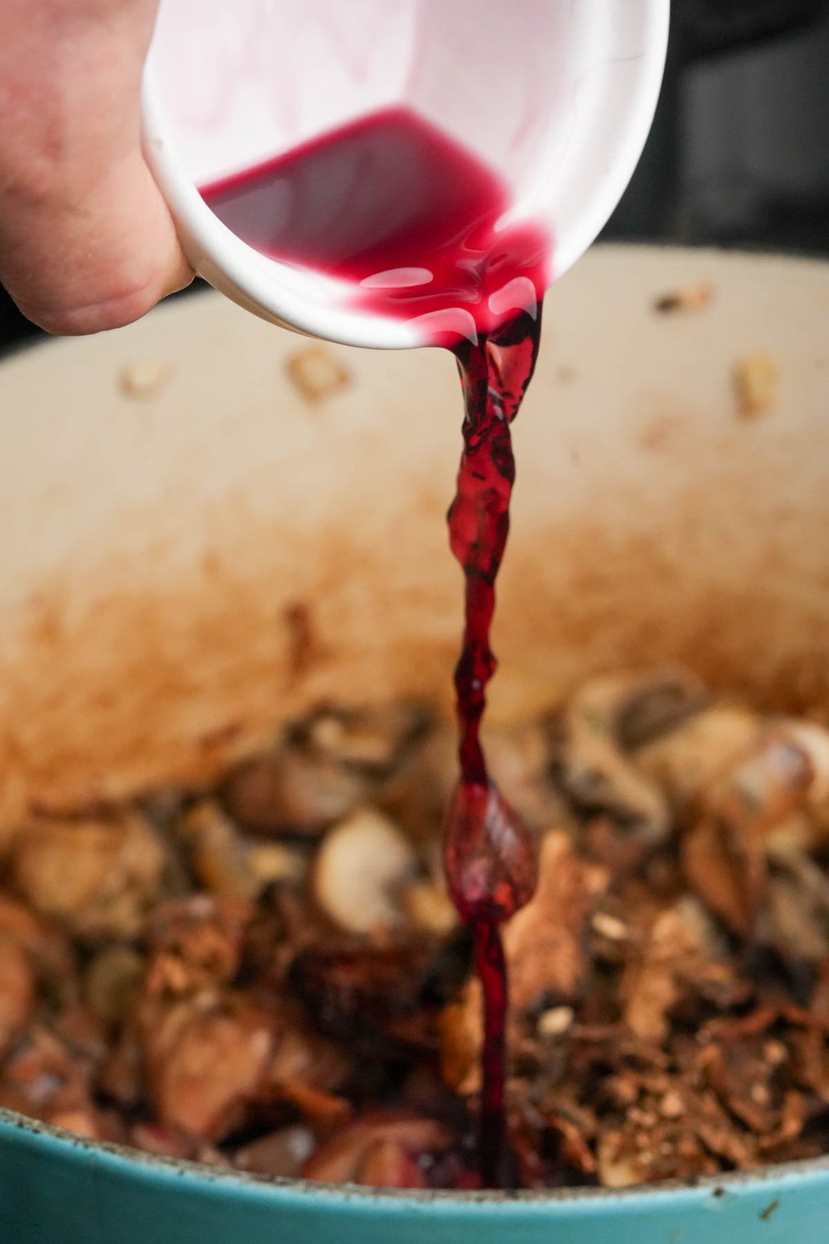 A close-up of red wine being poured from a white cup into a pot filled with sliced mushrooms and other ingredients.