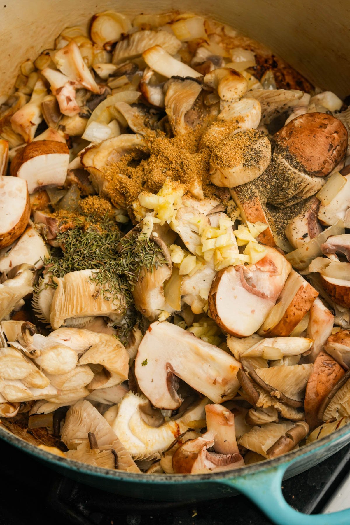 A close-up of assorted chopped mushrooms, garlic, and herbs in a large pot, ready to be cooked. The ingredients are uncooked and spread out, showing textures and natural colors.