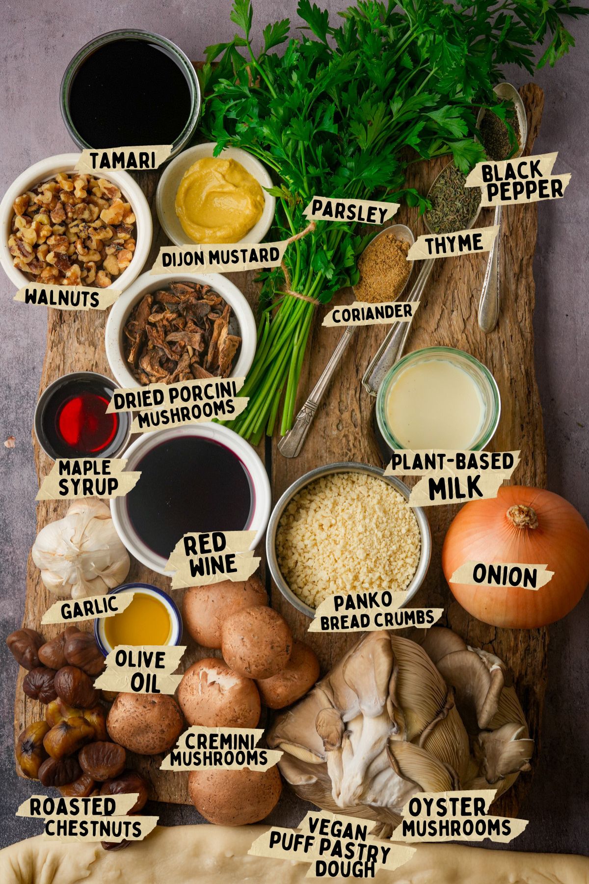 A wooden board with labeled ingredients for a vegan mushroom Wellington recipe, including herbs, nuts, mushrooms, onion, garlic, plant-based milk, olive oil, panko, vegan puff pastry dough, chestnuts, tamari, Dijon mustard, maple syrup, and red wine.