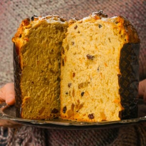A person holding a sliced panettone, an Italian sweet bread loaf filled with raisins and candied fruit, showing its soft, fluffy interior. The person is wearing a textured, multicolored sweater.