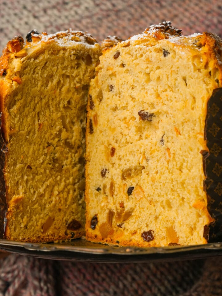 A person holding a sliced panettone, an Italian sweet bread loaf filled with raisins and candied fruit, showing its soft, fluffy interior. The person is wearing a textured, multicolored sweater.