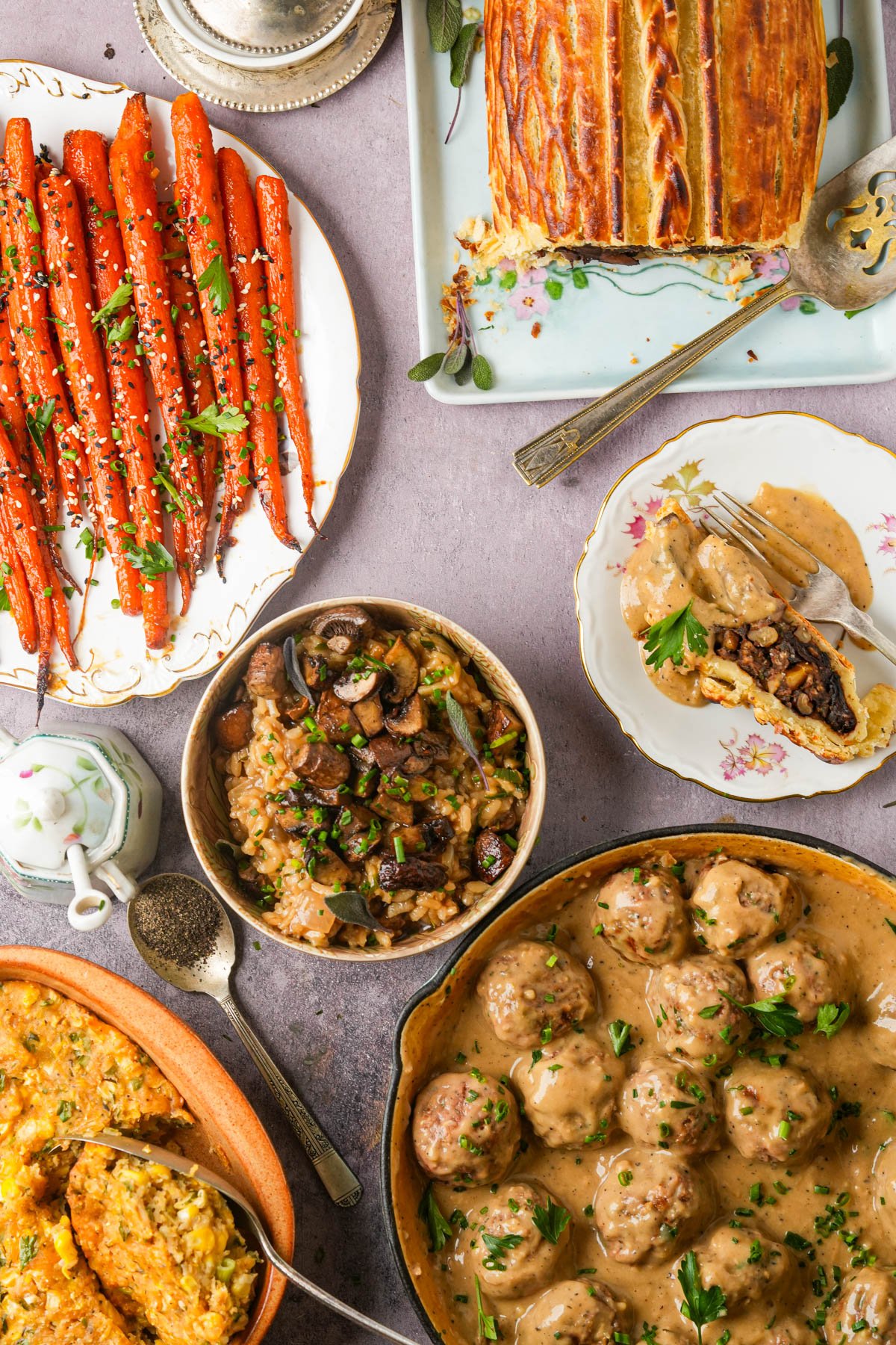A table with plates of maple roasted carrots, mushroom risotto, vegan mushroom Wellington, vegan corn casserole, vegan Swedish meatballs in sauce, and a cup of black pepper, all garnished with herbs on a light surface.