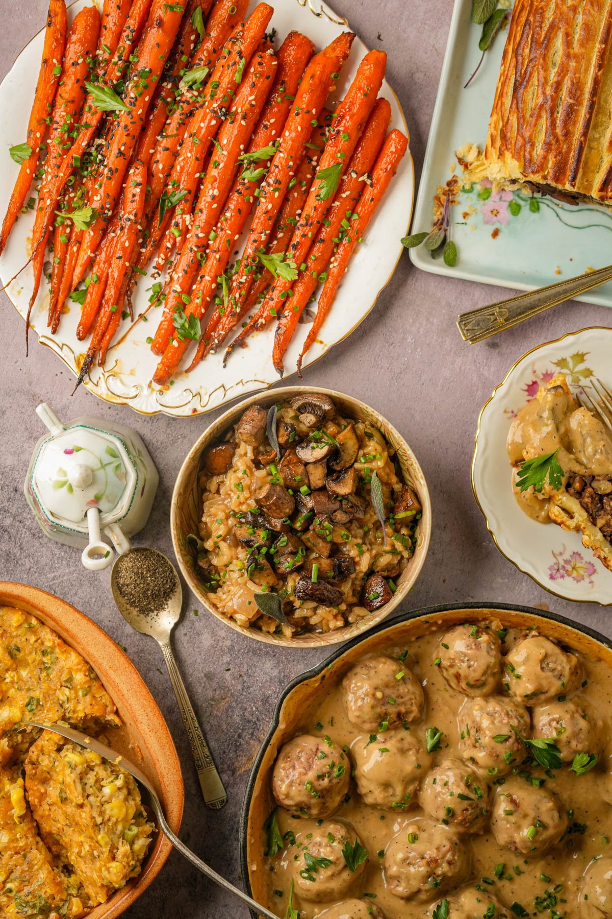 A table with plates of maple roasted carrots, mushroom risotto, vegan mushroom Wellington, vegan corn casserole, vegan Swedish meatballs in sauce, and a cup of black pepper, all garnished with herbs on a light surface.