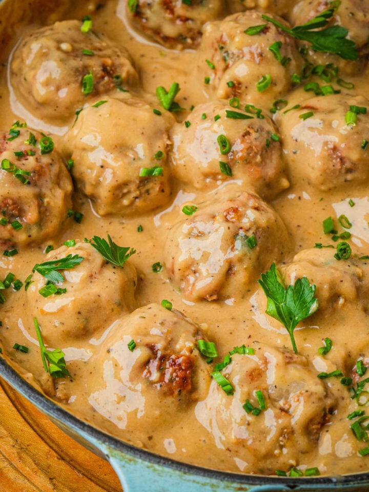 Vegan Swedish meatballs coated in creamy brown gravy, garnished with chopped parsley and chives, in a skillet on a wooden surface.