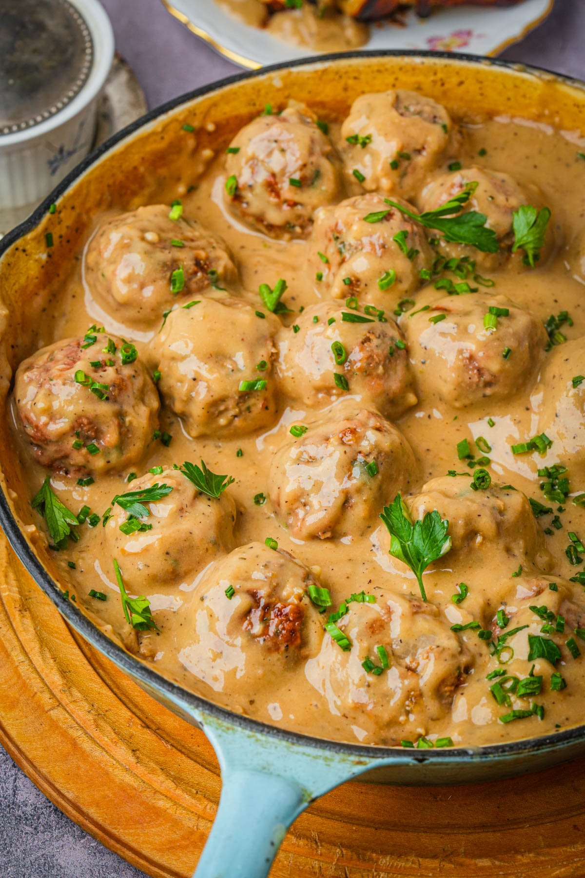 A skillet filled with vegan Swedish meatballs covered in creamy brown sauce, garnished with chopped parsley and chives, sits on a wooden surface.