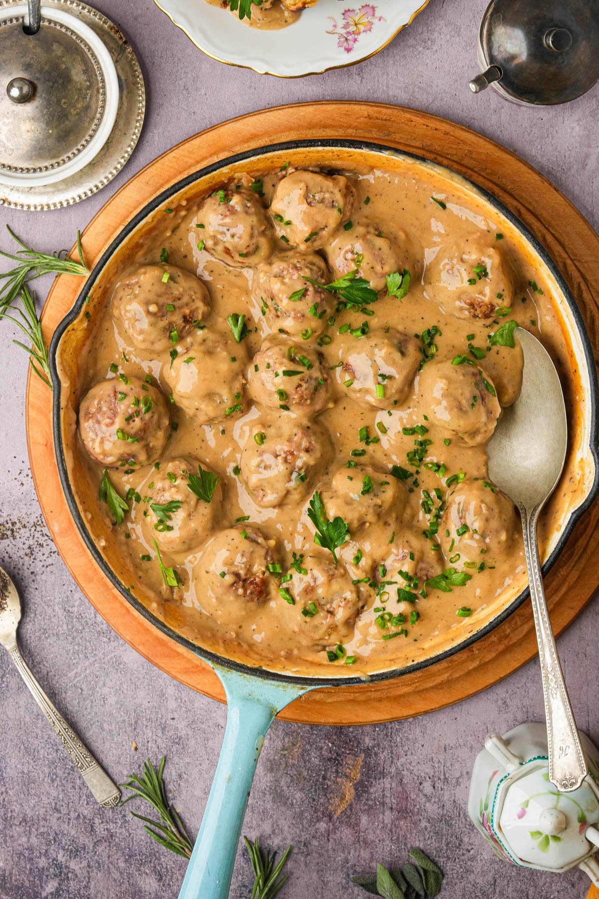 A skillet filled with vegan Swedish meatballs covered in creamy brown gravy, garnished with chopped parsley, sits on a wooden board. A large spoon rests in the skillet, with herbs and utensils arranged around it.
