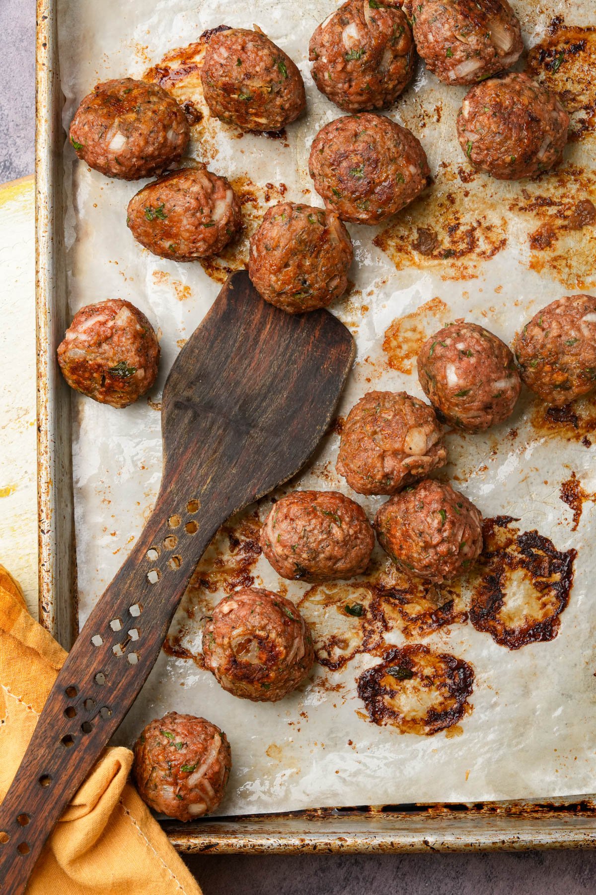 A baking sheet with browned vegan Swedish meatballs on parchment paper, some scattered and some grouped. A dark wooden spatula and a yellow cloth are placed on the left side of the tray.