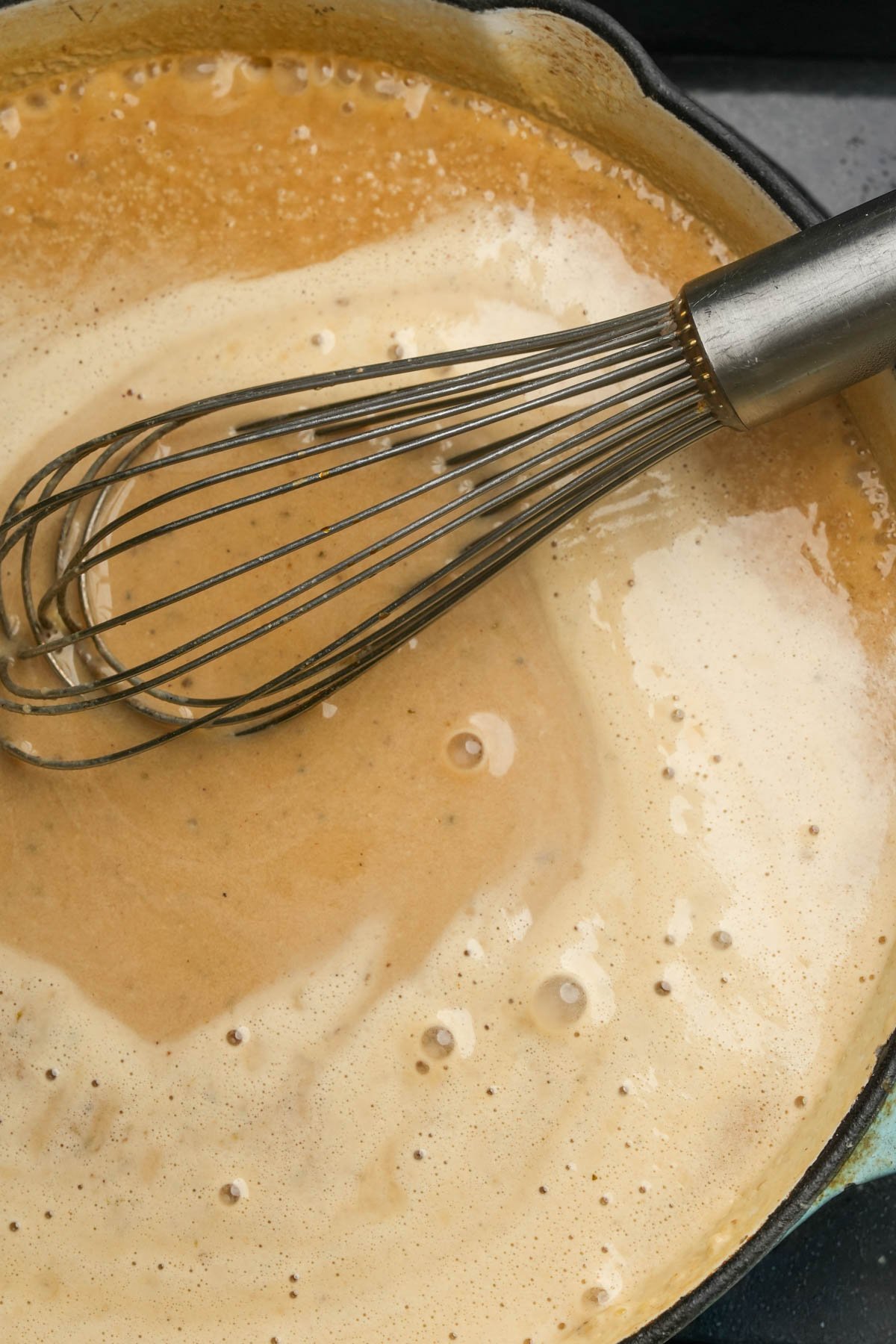 A metal whisk rests in a pan of creamy, light brown vegan Swedish meatball gravy.