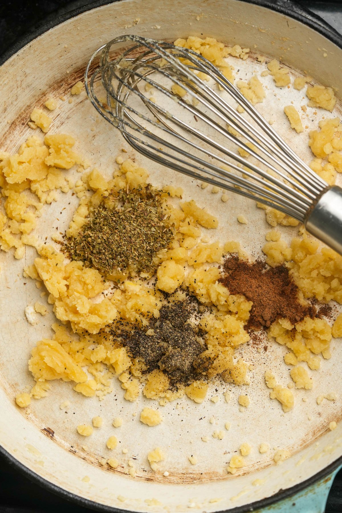 A metal whisk rests in a pan with yellow roux and piles of dried herbs, ground black pepper, and ground nutmeg, ready to be mixed together.