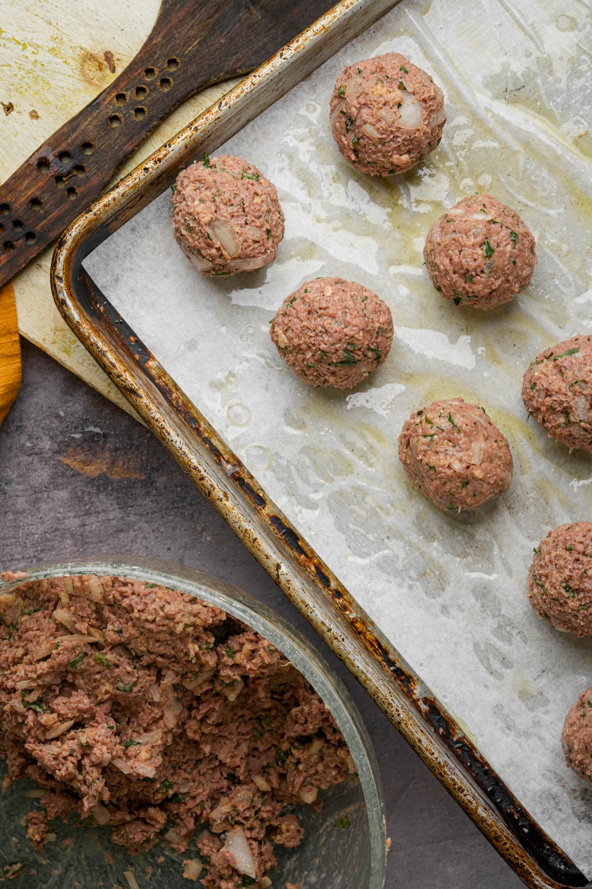 Raw vegan Swedish meatballs with chopped onions and herbs are placed on a parchment-lined baking sheet. A bowl of extra meatball mixture and a wooden spatula sit nearby on a textured countertop.