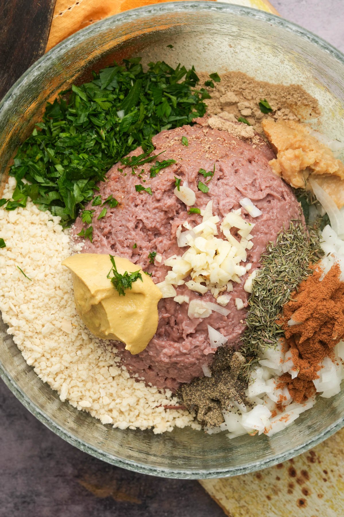 A glass bowl filled with ground beyond meat, chopped parsley, breadcrumbs, mustard, minced garlic, chopped onions, and various spices, all arranged separately and ready to be mixed to be formed into vegan Swedish meatballs.