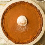 A whole vegan sweet potato pie with a slightly browned crust, topped with a dollop of whipped cream and a sprinkle of cinnamon, on a light-colored plate.