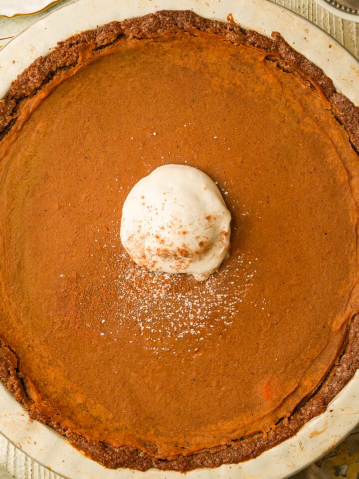 A whole vegan sweet potato pie with a slightly browned crust, topped with a dollop of whipped cream and a sprinkle of cinnamon, on a light-colored plate.