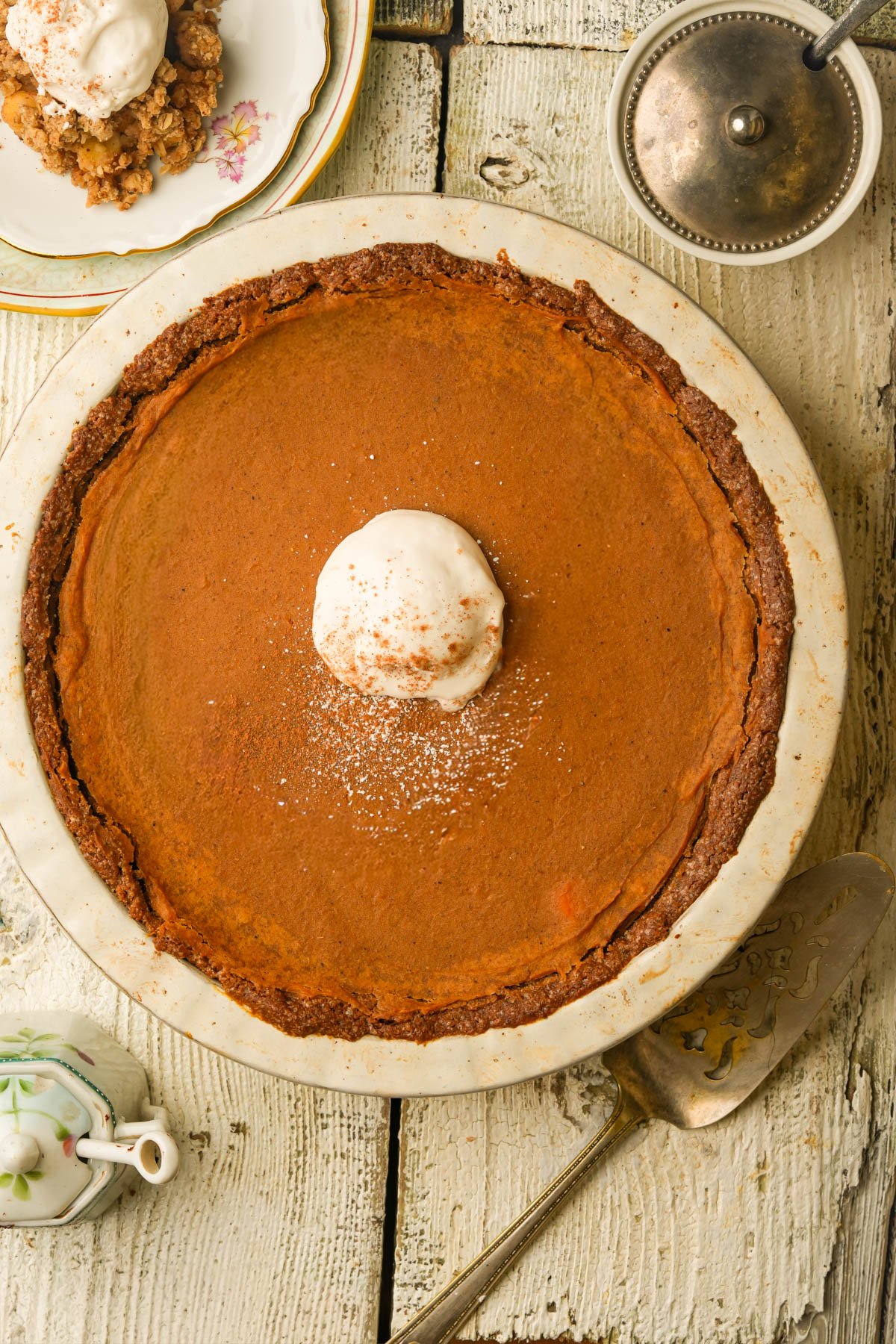 A pumpkin pie topped with a dollop of whipped cream and a dusting of cinnamon sits on a rustic white wooden table, with a pie server, teapot, and a plate with a crumble dessert nearby.