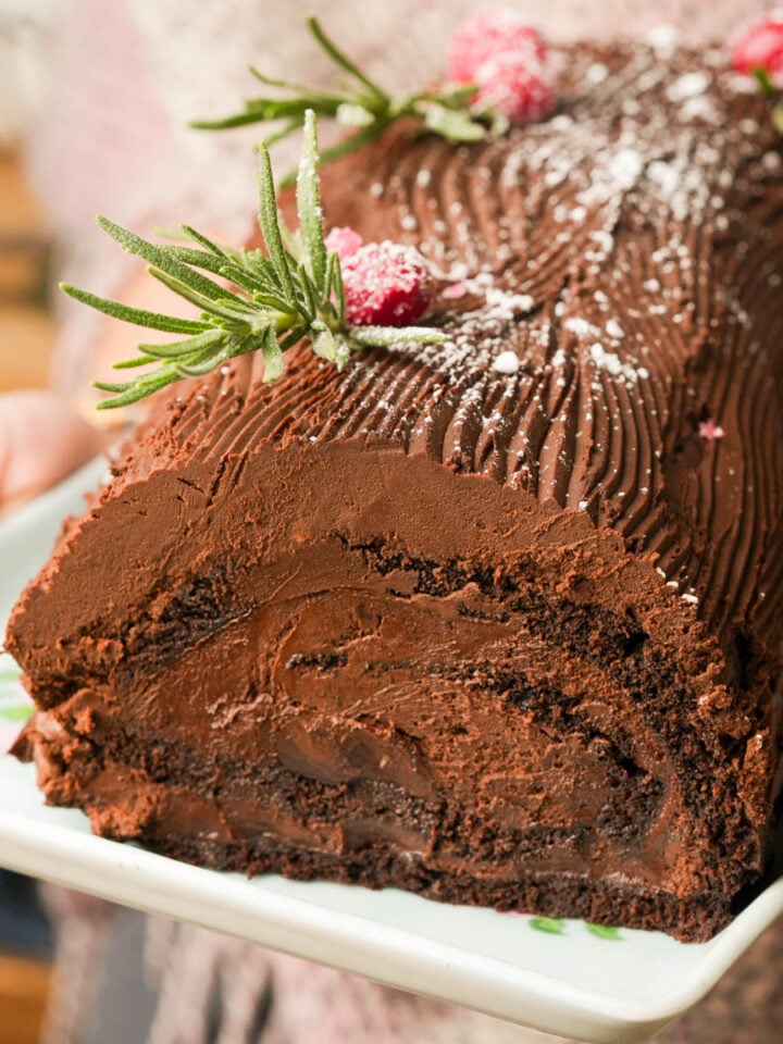 A person holds a platter with a chocolate yule log cake decorated with chocolate frosting, sugared cranberries, and sprigs of rosemary, dusted with powdered sugar.