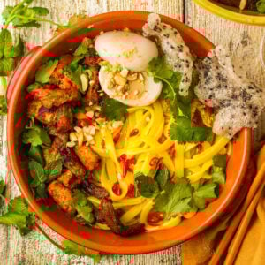 A bowl of yellow vegan mi quang noodles topped with grilled tofu and soy curl, fresh herbs, chopped peanuts, red chili slices, a halved vegan boiled egg, and crispy crackers, served on a rustic wooden table.