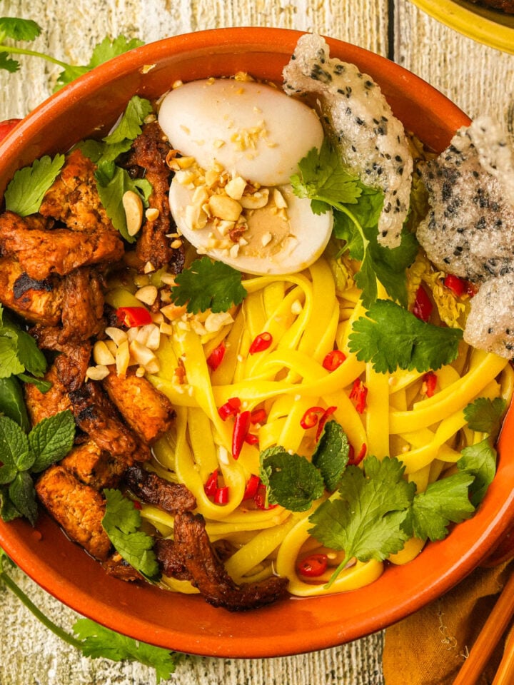 A bowl of yellow vegan mi quang noodles topped with grilled tofu and soy curl, fresh herbs, chopped peanuts, red chili slices, a halved vegan boiled egg, and crispy crackers, served on a rustic wooden table.