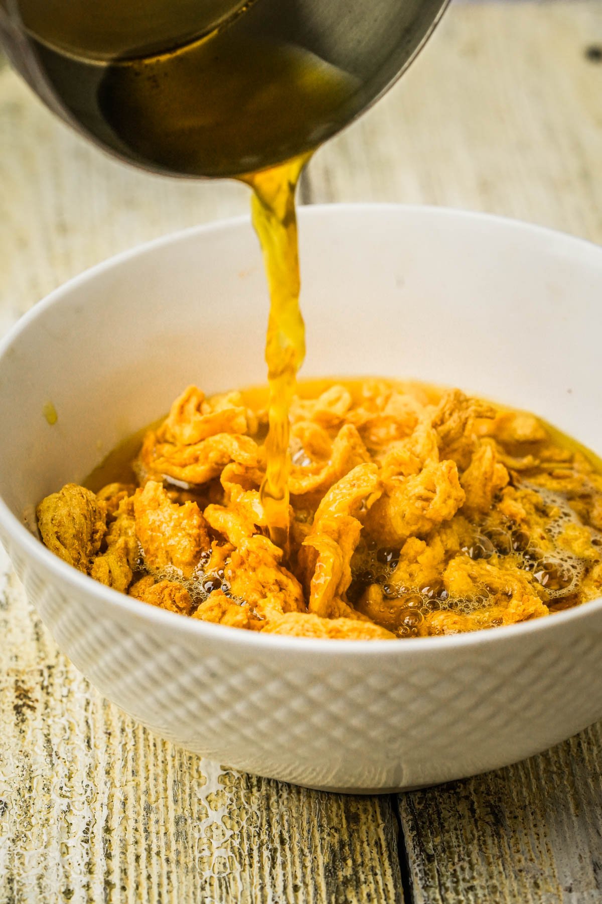 Golden broth is being poured from a metal pot into a white bowl filled with soy curls, all set on a rustic wooden surface.