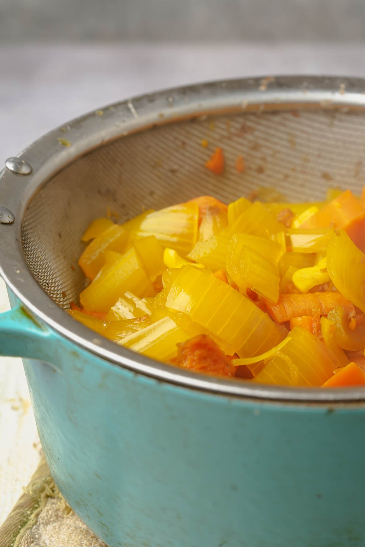A metal strainer filled with cooked yellow onions, carrots, and other vegetables rests on top of a turquoise pot. The vegetables are being strained to leave behind vegan mi quang stock.