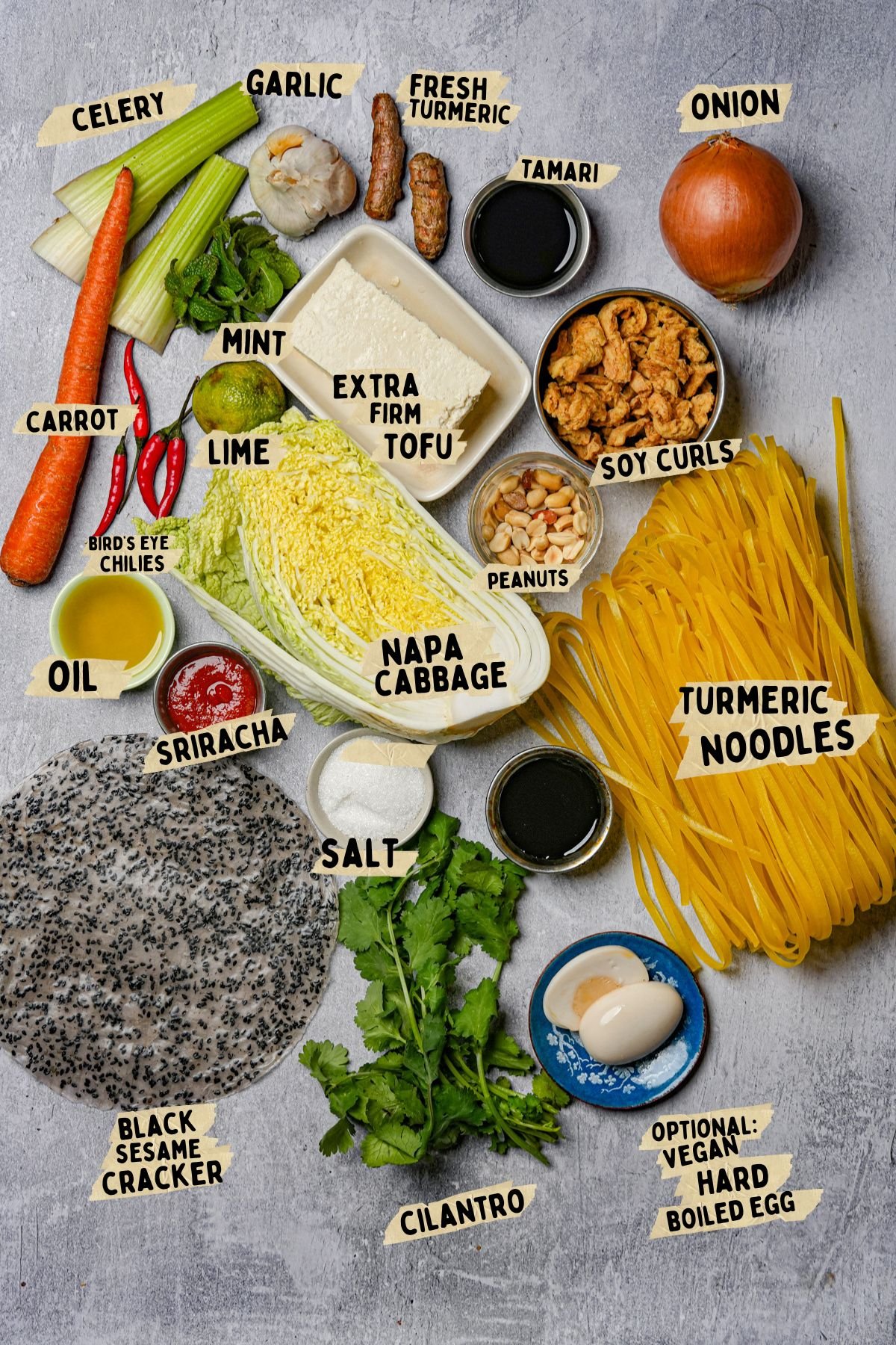 A flat lay of vegan noodle bowl ingredients, including turmeric noodles, napa cabbage, extra firm tofu, soy curls, black sesame crackers, carrot, celery, onion, garlic, fresh turmeric, mint, cilantro, peanuts, chilies, oil, tamari, sriracha, and salt.