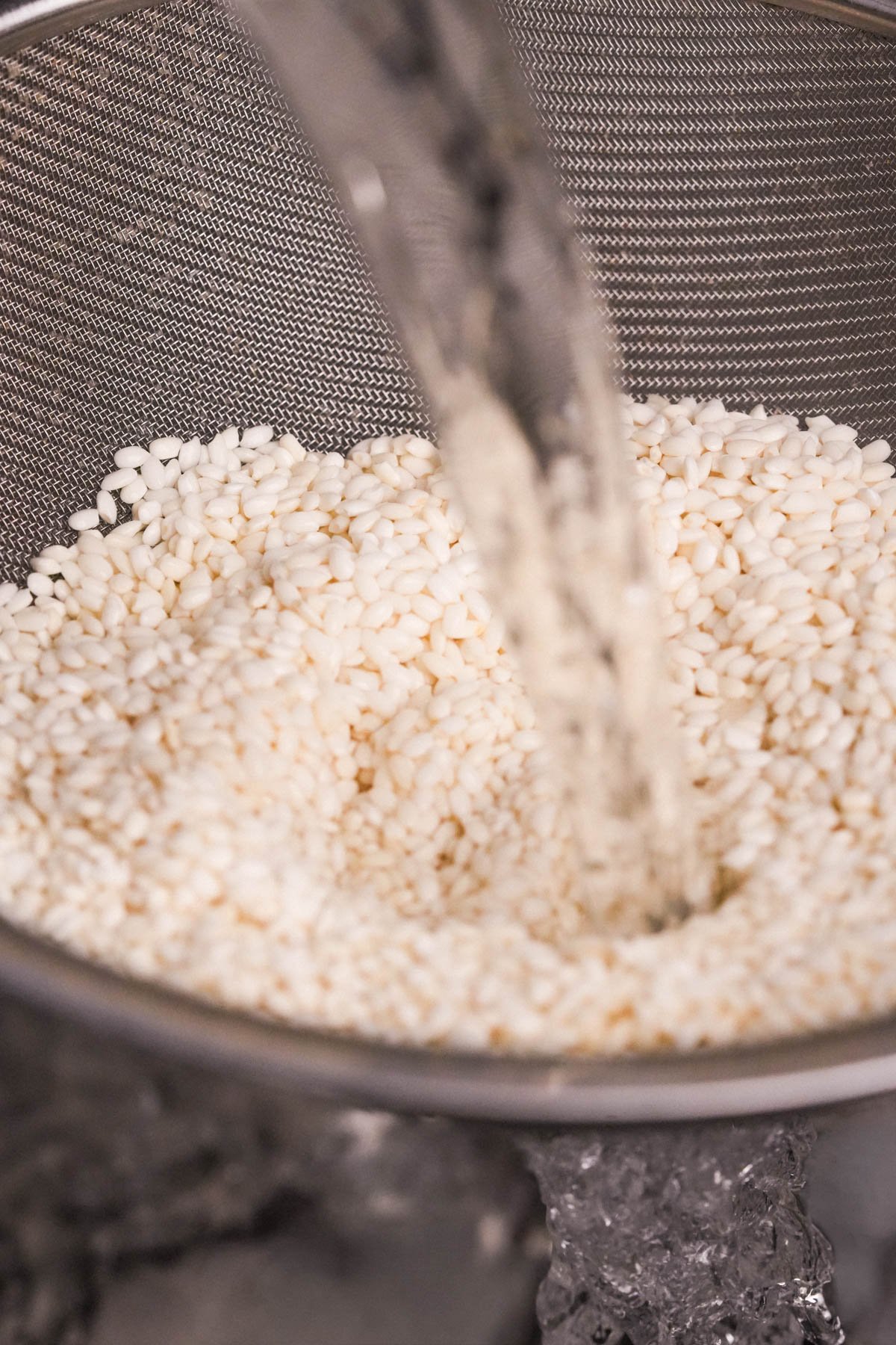 Water is being poured over a pile of Vietnamese sticky rice grains in a metal mesh strainer, likely to rinse or wash the rice before cooking.