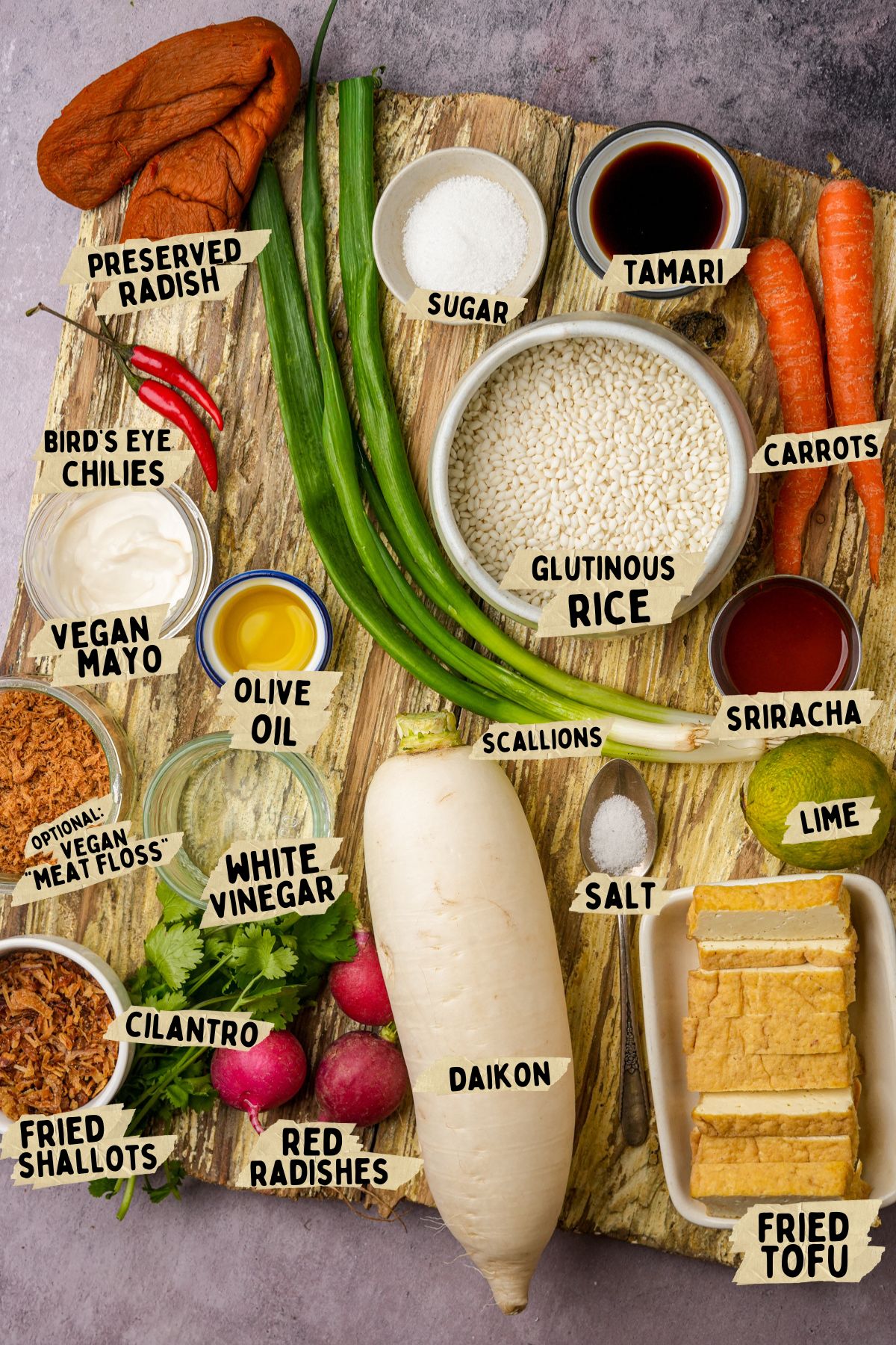 A variety of labeled vegan ingredients for Vietnamese Sticky Rice on a wooden board, including glutinous rice, preserved radish, scallions, daikon, carrots, fried tofu, vegan mayo, chilies, lime, radishes, condiments, and seasonings.