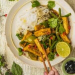 A hand holds chopsticks picking up a piece of lemongrass tofu from a plate with stir-fried tofu, green beans, red chili, herbs, rice noodles, and half a lime, garnished with cilantro on a rustic table.