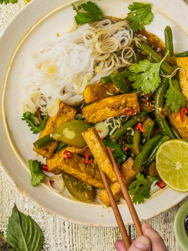 A hand holds chopsticks picking up a piece of lemongrass tofu from a plate with stir-fried tofu, green beans, red chili, herbs, rice noodles, and half a lime, garnished with cilantro on a rustic table.