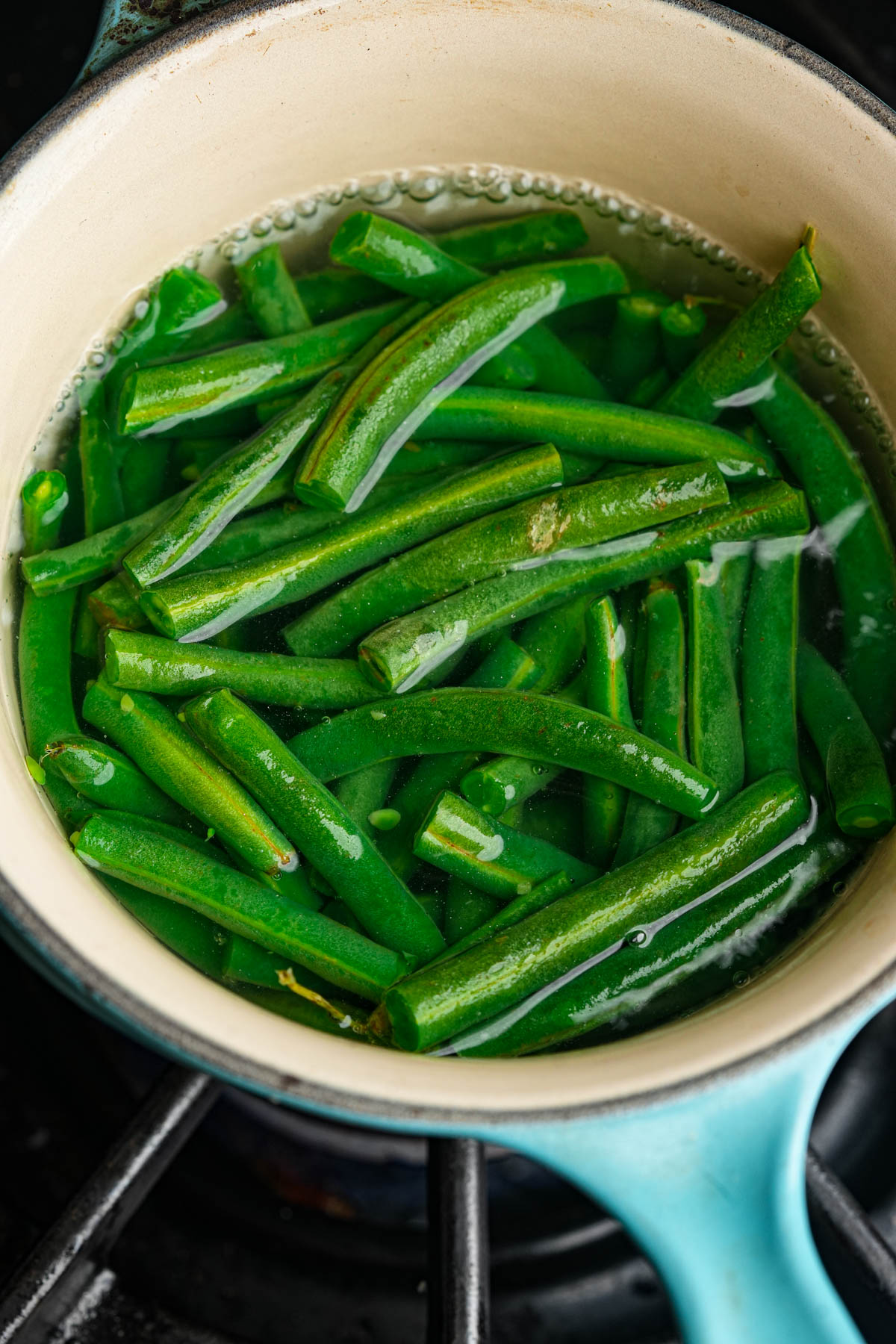 Fresh green beans being boiled in water inside a light-colored pot on a stovetop. The beans are bright green and partially submerged in bubbling water.