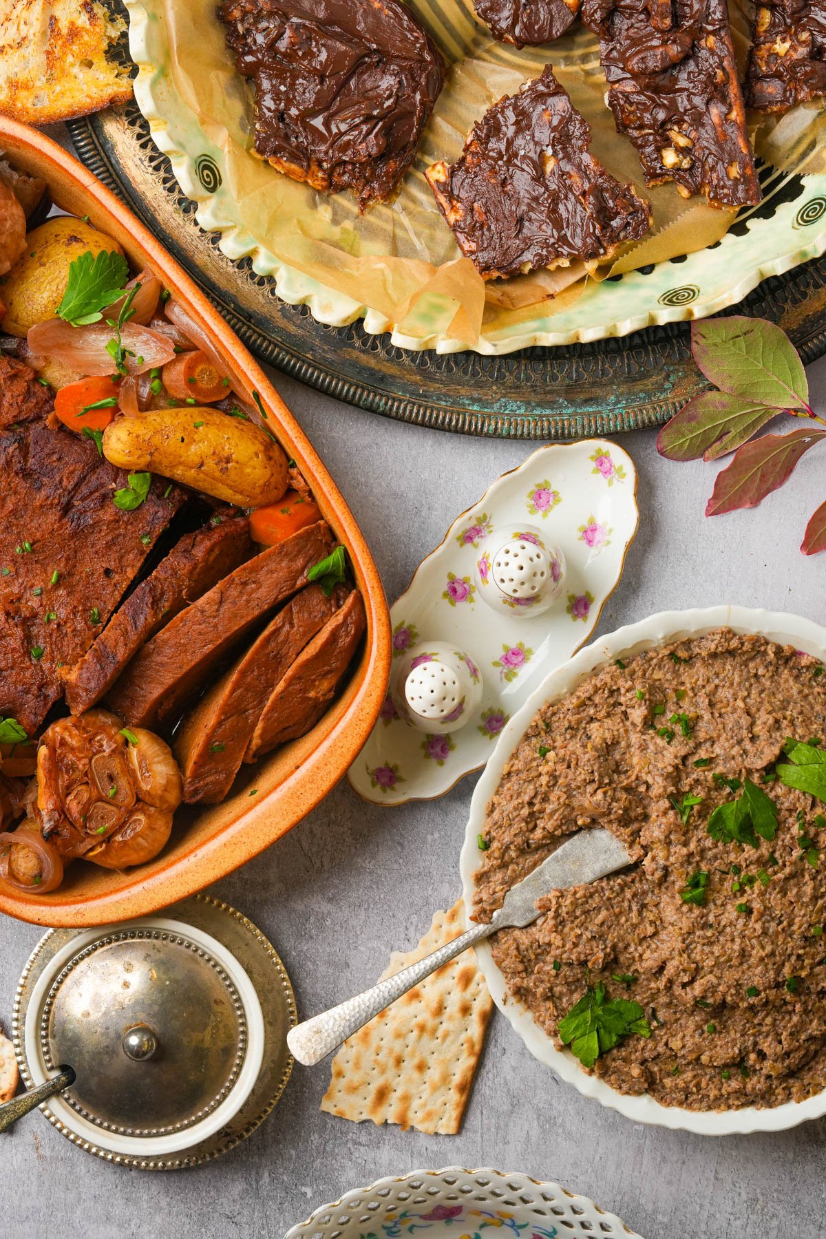 A table with vegan Jewish holiday foods: chocolate-covered matzo tofee, a dish of Jewish vegan brisket with vegetables, a plate of vegan chopped liver on a gray surface.