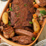 A baked Jewish vegan brisket in a dish with whole potatoes, carrots, onions, and halved heads of garlic, garnished with chopped parsley.