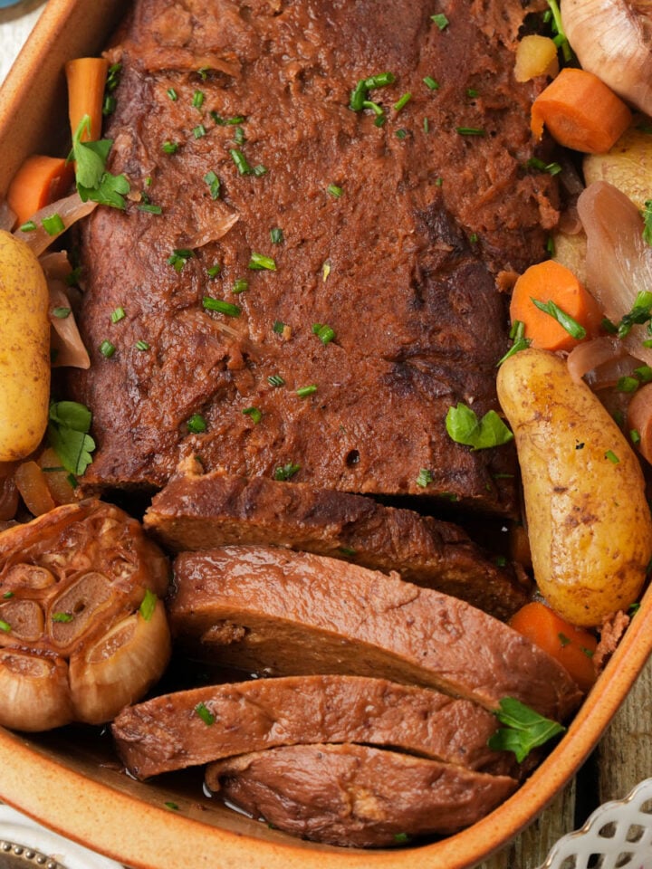 A baked Jewish vegan brisket in a dish with whole potatoes, carrots, onions, and halved heads of garlic, garnished with chopped parsley.