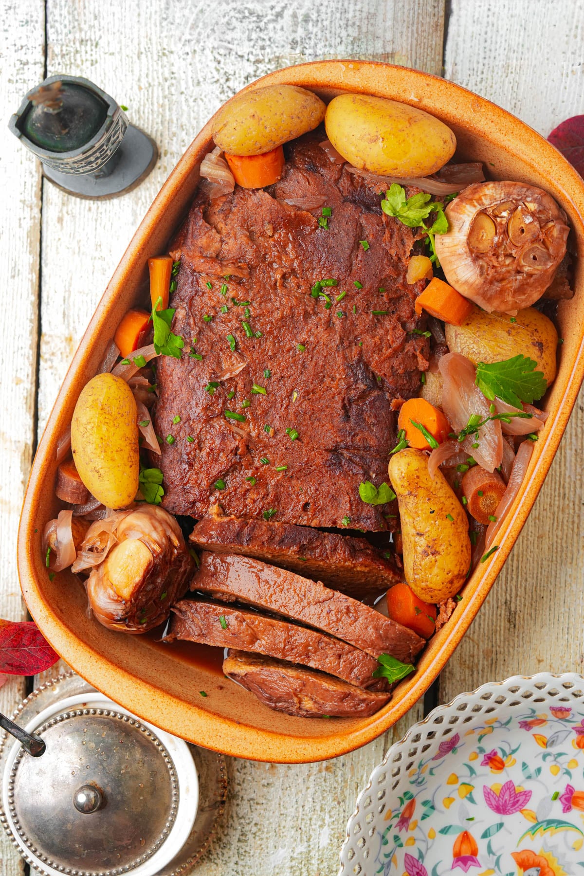 A large piece of Jewish vegan brisket, sliced at one end, surrounded by golden potatoes, carrots, onions, and whole roasted garlic bulbs, garnished with chopped parsley on a white wooden table.