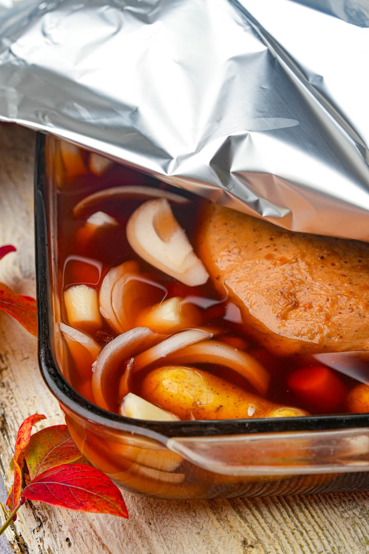 A glass baking dish filled with sliced onions, potatoes, and a piece of Jewish vegan brisket in a reddish broth, partially covered with aluminum foil, sits on a light wooden surface near red autumn leaves.