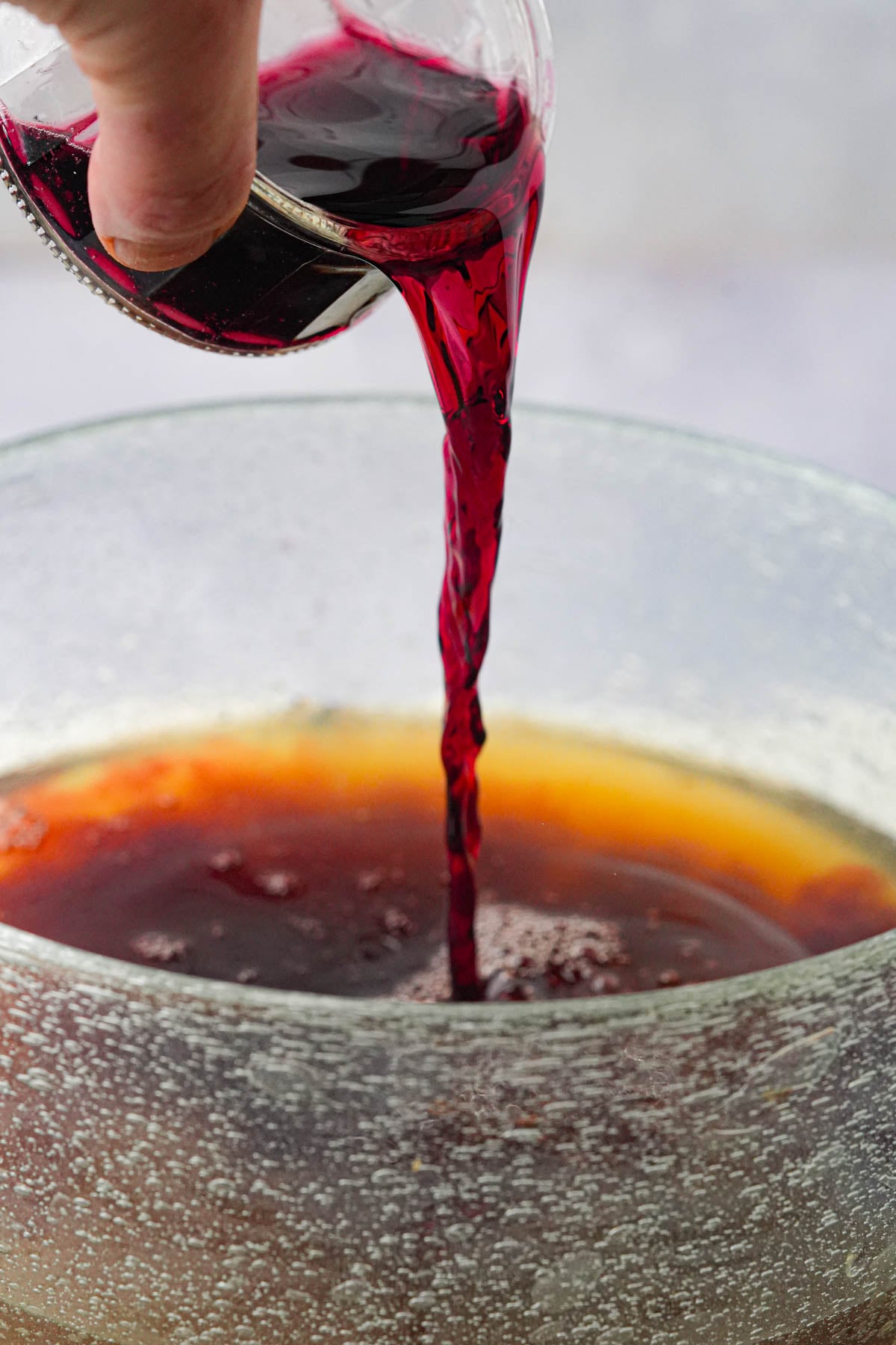 A hand pours red wine from a small glass into a large bowl filled with vegetable stock and tamari, with condensation visible on the bowl's surface.