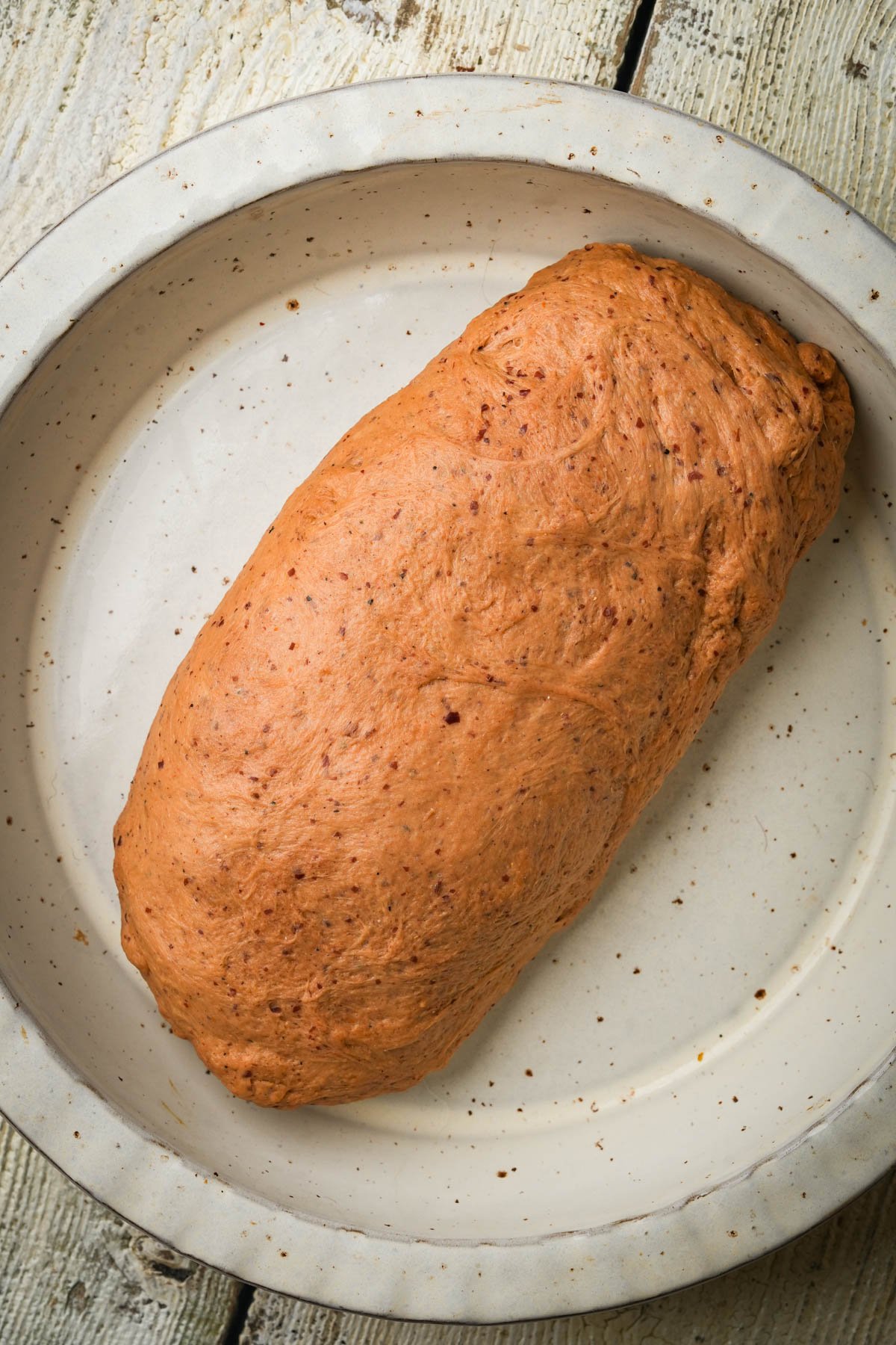 A loaf-shaped mound of Jewish vegan brisket dough sits in a round, off-white ceramic dish on a rustic wooden surface.