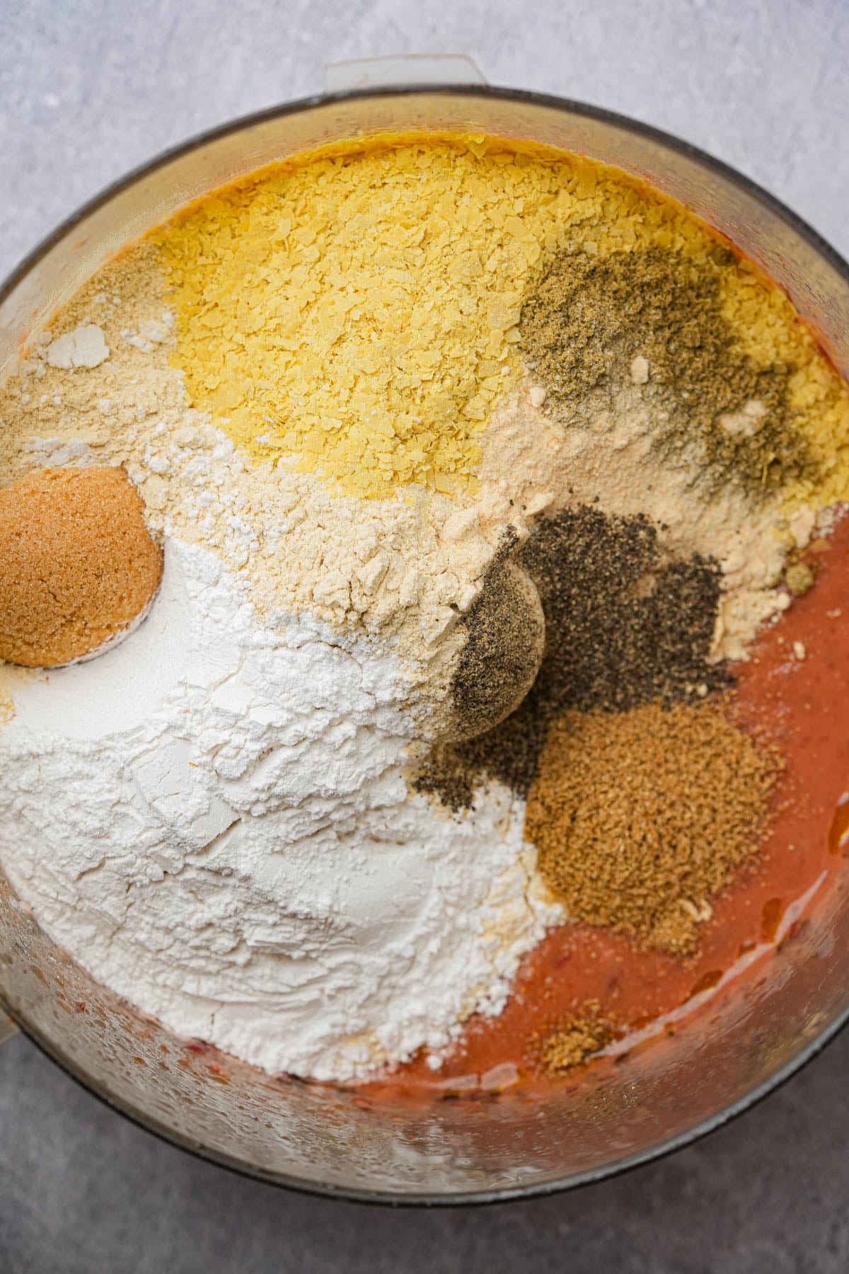 A close-up of a food processor bowl filled with various powders and spices, including flour, nutritional yeast, black pepper, and other seasonings, arranged in distinct, colorful piles.