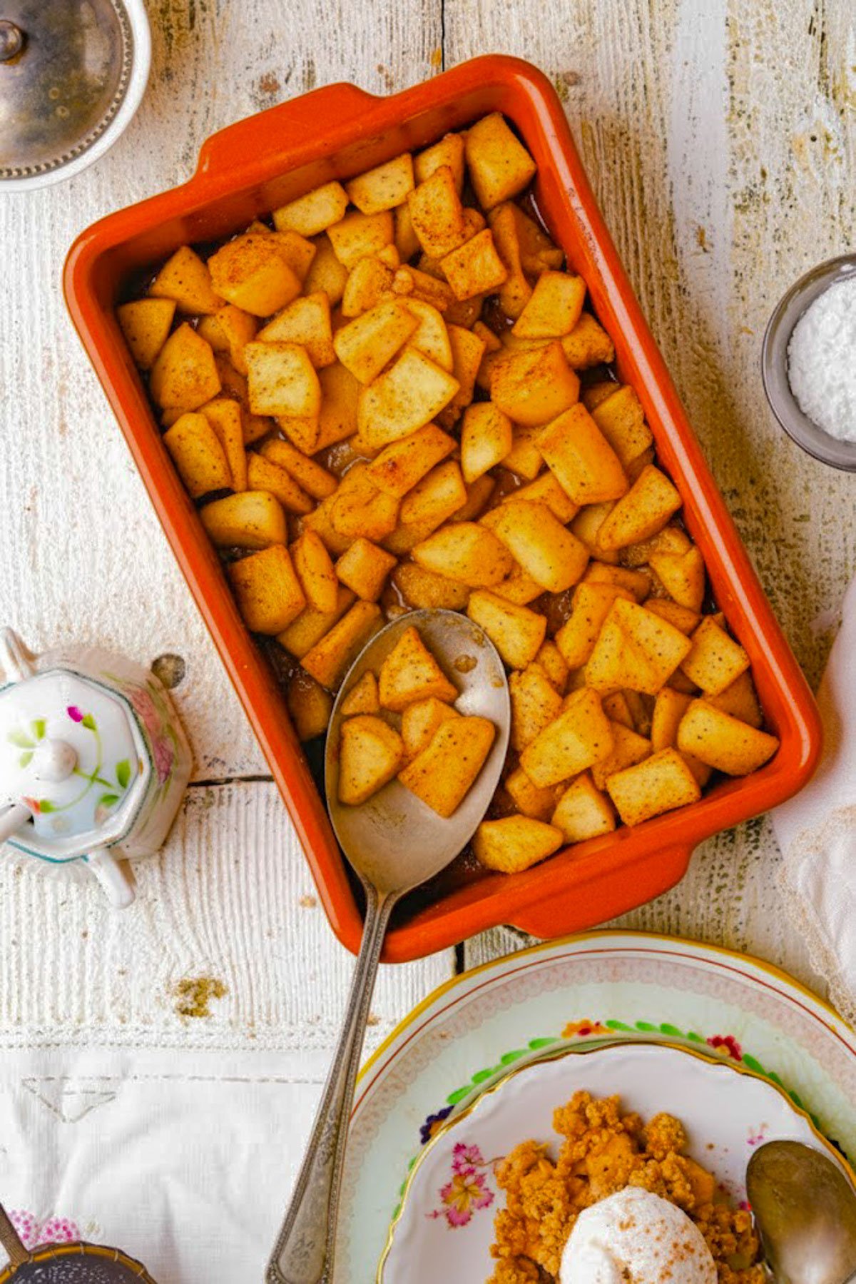 A rectangular orange baking dish filled with diced, baked apples coated in cinnamon, served as a Vegan Apple Crisp. A large serving spoon rests inside, and the rustic white table is surrounded by plates and a sugar bowl.