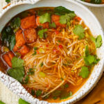 A bowl of spicy vegan bun bo hue noodle soup with tofu, leafy greens, fresh cilantro, and red chili slices in a clear, flavorful broth. The noodles are surrounded by vegetables and herbs.