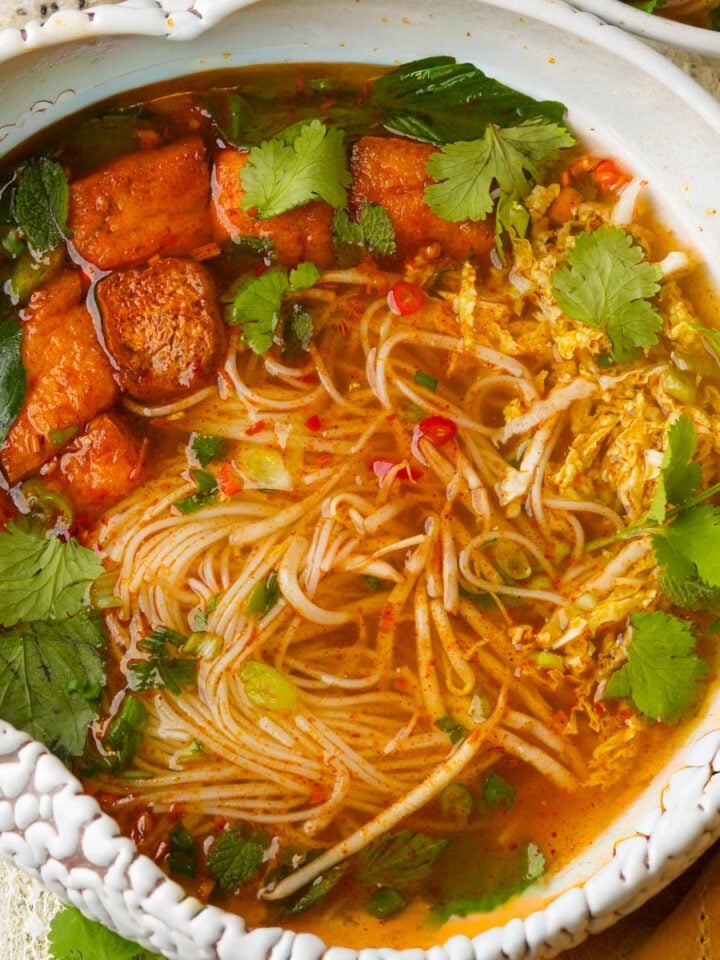 A bowl of spicy vegan bun bo hue noodle soup with tofu, leafy greens, fresh cilantro, and red chili slices in a clear, flavorful broth. The noodles are surrounded by vegetables and herbs.