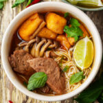 A bowl of vegan bun rieu noodle soup with beef slices, fried tofu, mushrooms, fresh herbs, and a wedge of lime, served in a white bowl on a rustic wooden table.
