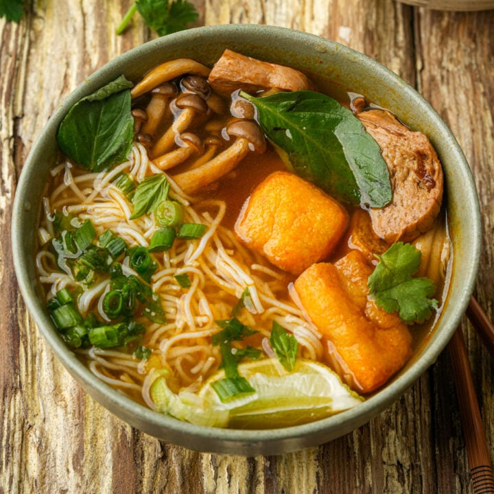 A bowl of vegan bun rieu noodle soup with mushrooms, tofu, sliced vegan ham, fresh herbs, lime wedge, and green onions in a flavorful broth, served on a rustic wooden table.