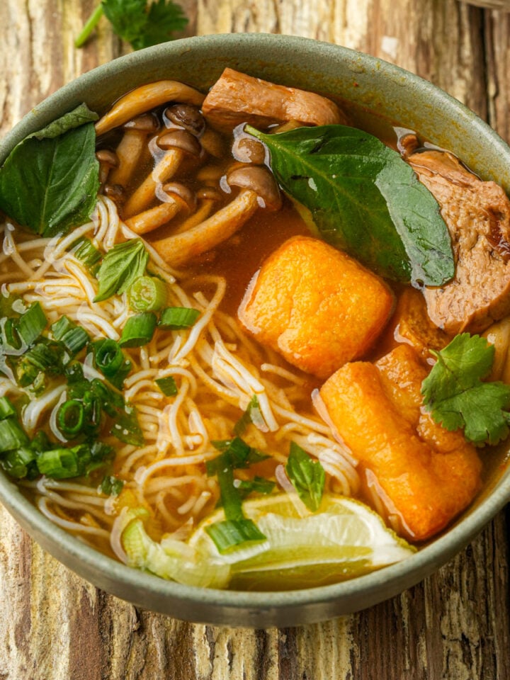 A bowl of vegan bun rieu noodle soup with mushrooms, tofu, sliced vegan ham, fresh herbs, lime wedge, and green onions in a flavorful broth, served on a rustic wooden table.